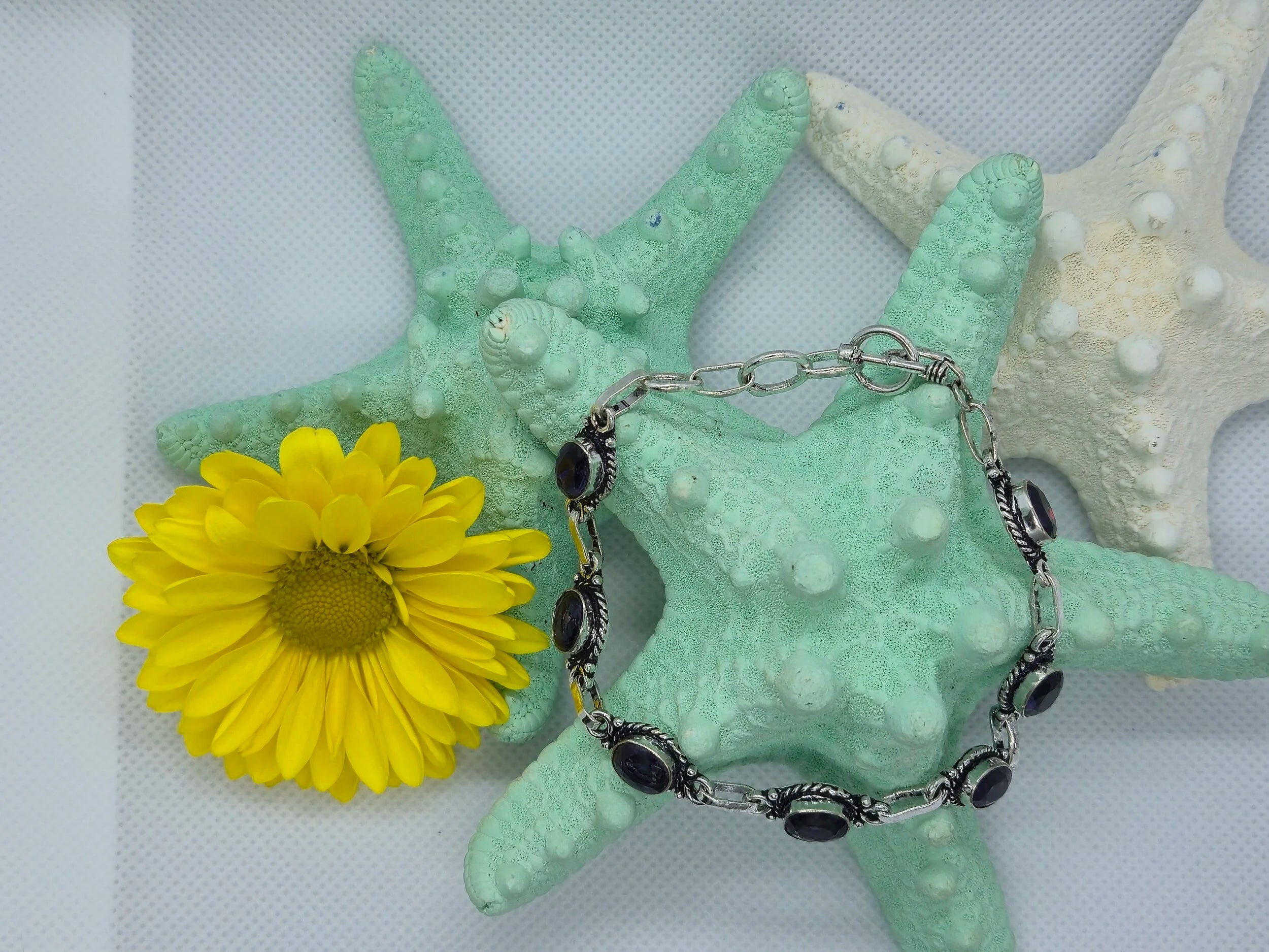 One Artisan Amethyst Silver Toggle Bracelet wrapped around an aquamarine starfish with another aquamarine starfish and white starfish in the background along with a sunflower. 