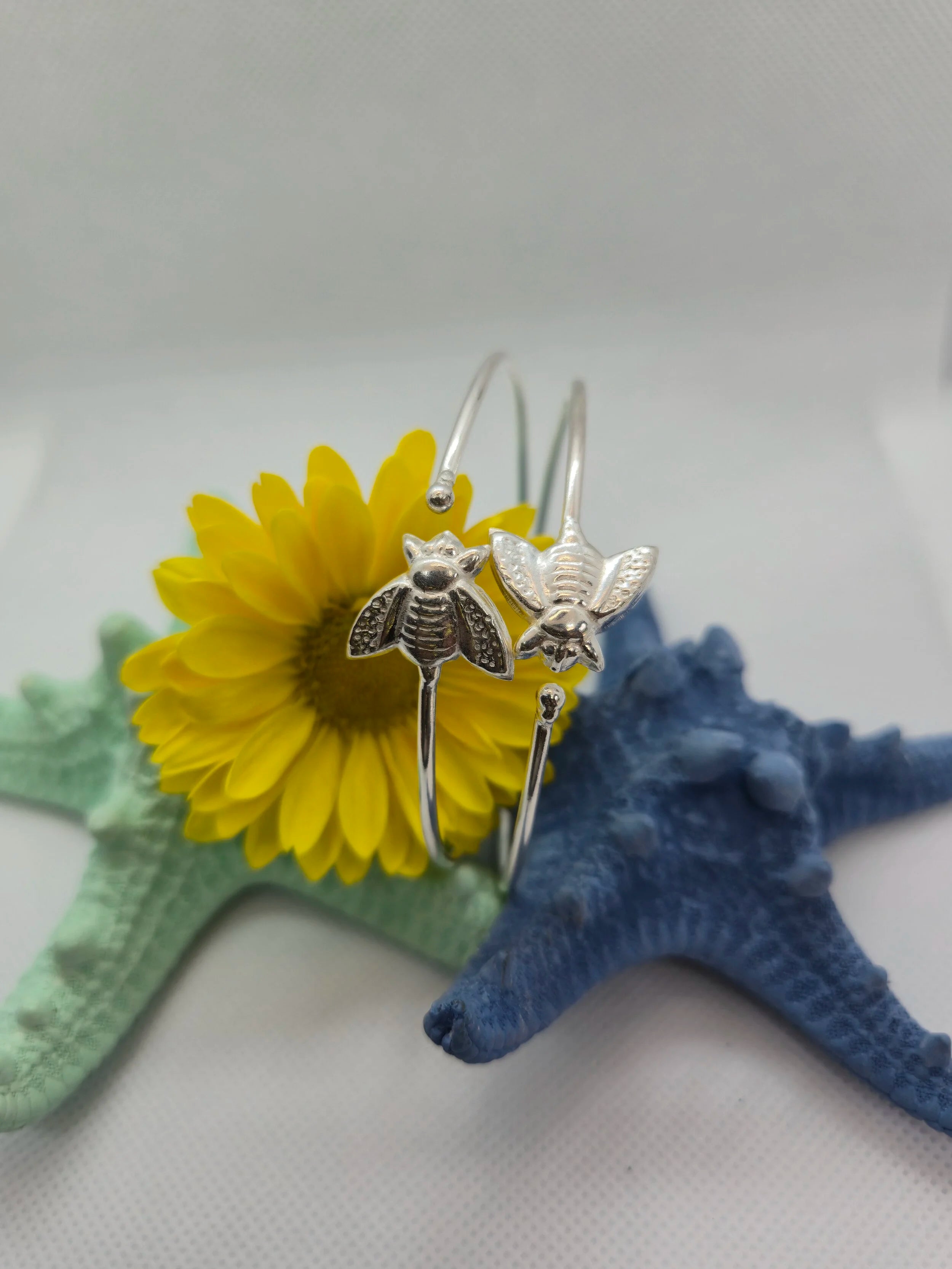 Two Artisan Silver Bumble Bee Bangles suspended over an aquamarine and navy starfish with a sunflower over a white background.