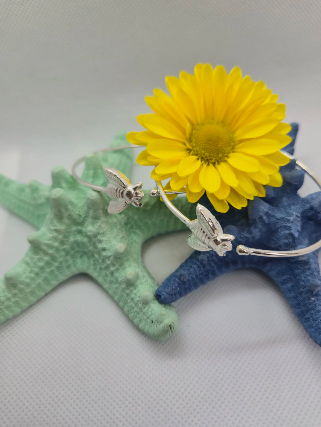 Two Artisan Silver Bumble Bee Bangles laid over an aquamarine and navy starfish with a sunflower on top.