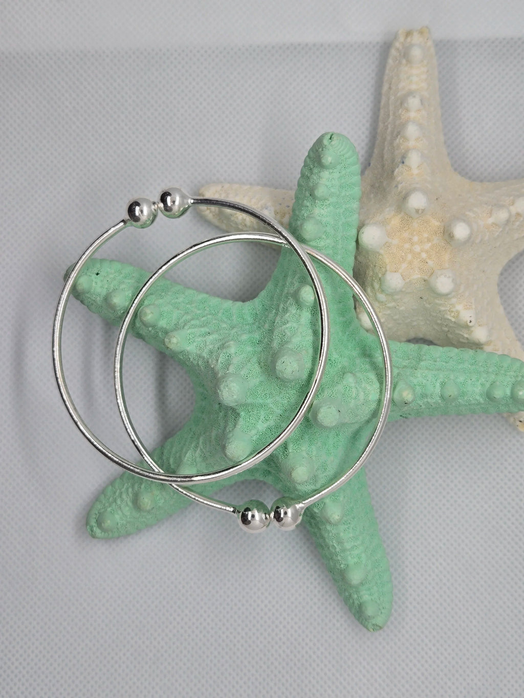 Two Heavy Silver West Indian Bangles resting on top of a aquamarine and white starfish with a white cloth background. 