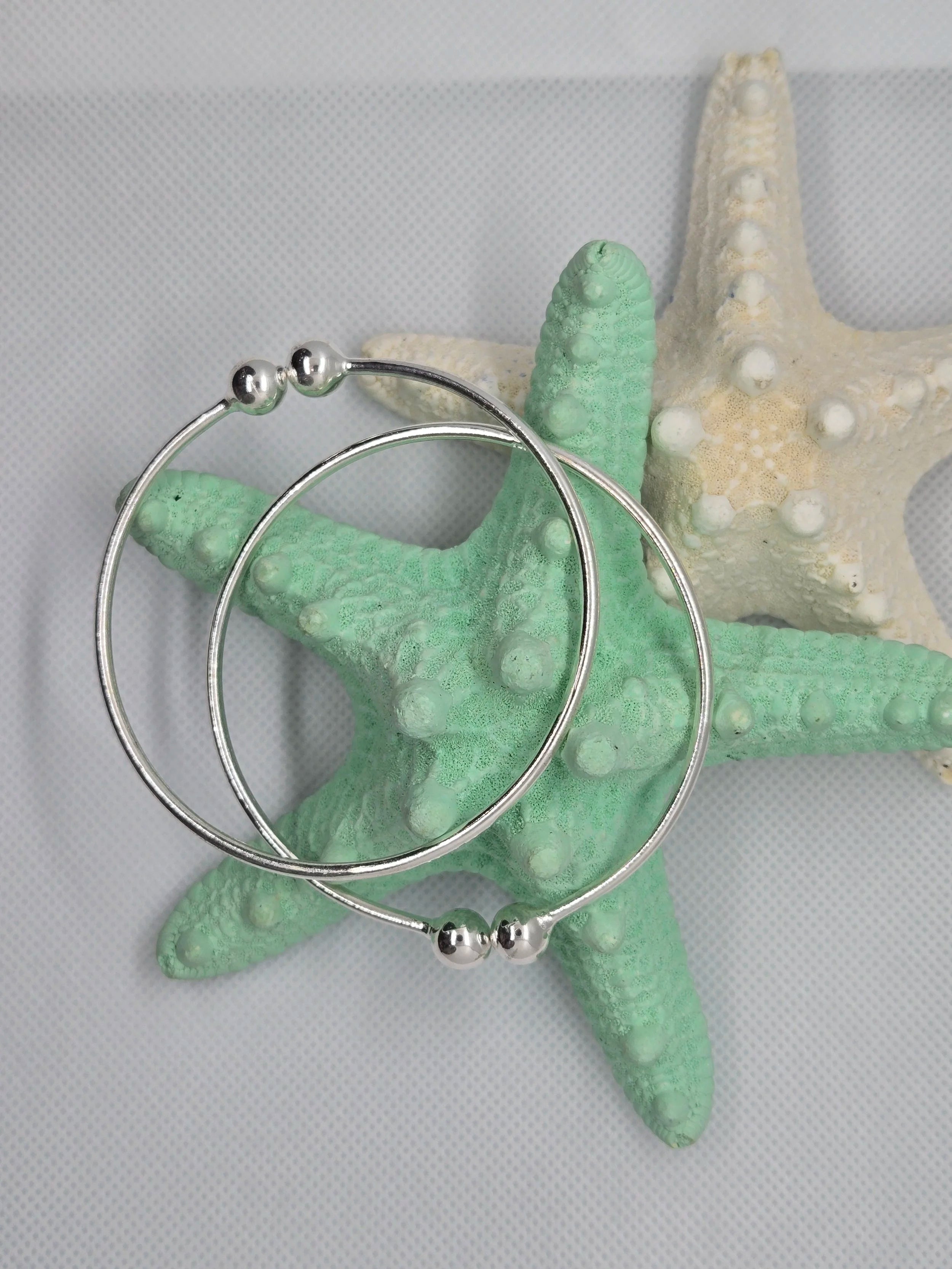 Two Heavy Silver West Indian Bangles resting on top of a aquamarine and white starfish with a white cloth background. 