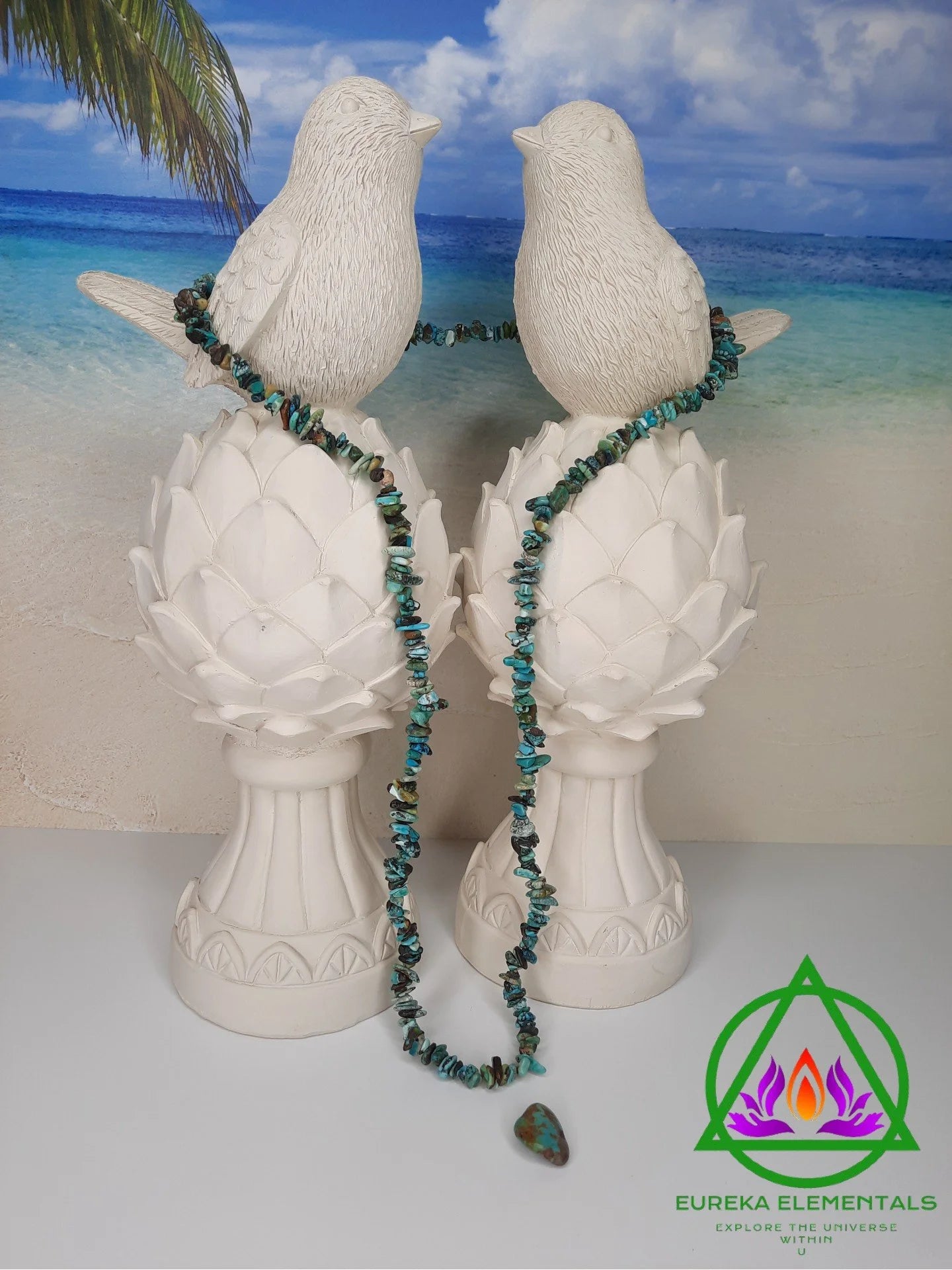 Hubei Turquoise Necklace and Crystal resting between two white dove statues with a beach backdrop.