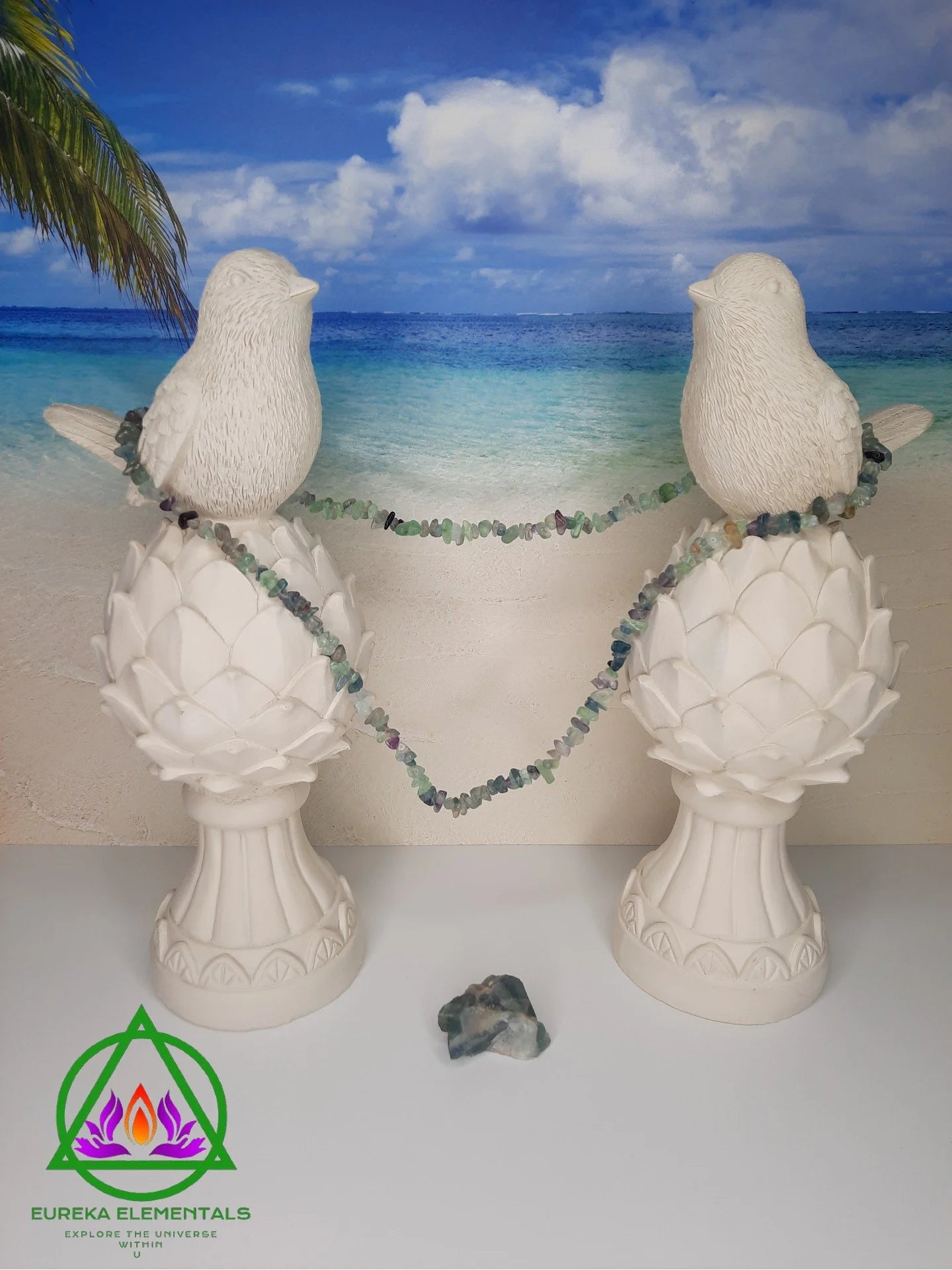 Rainbow Fluorite Necklace and Crystal resting between two white dove statues with a beach backdrop.