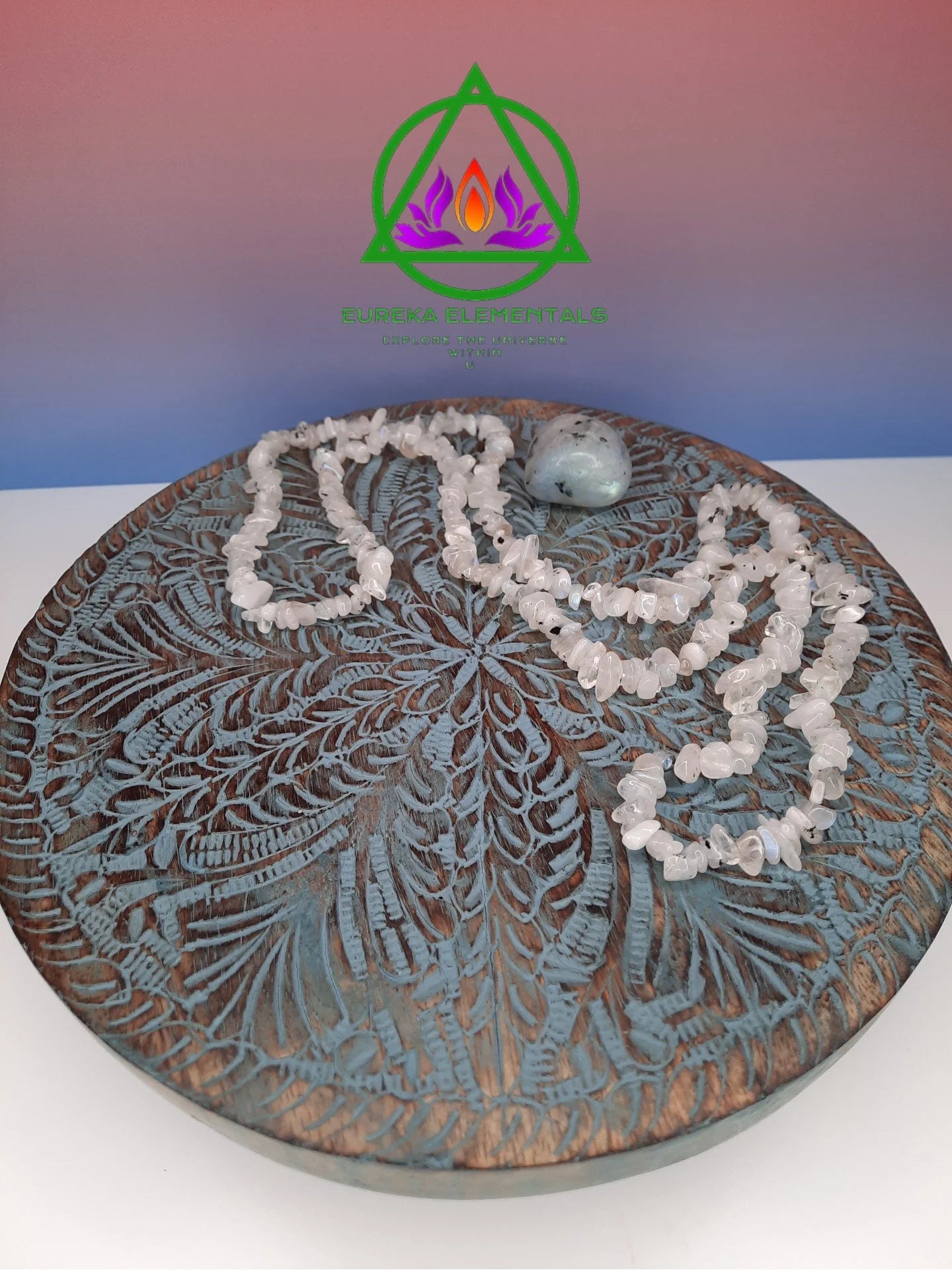 Rainbow Moonstone Necklace and Crystal resting on a wooden stand with a red and blue ombre backdrop.
