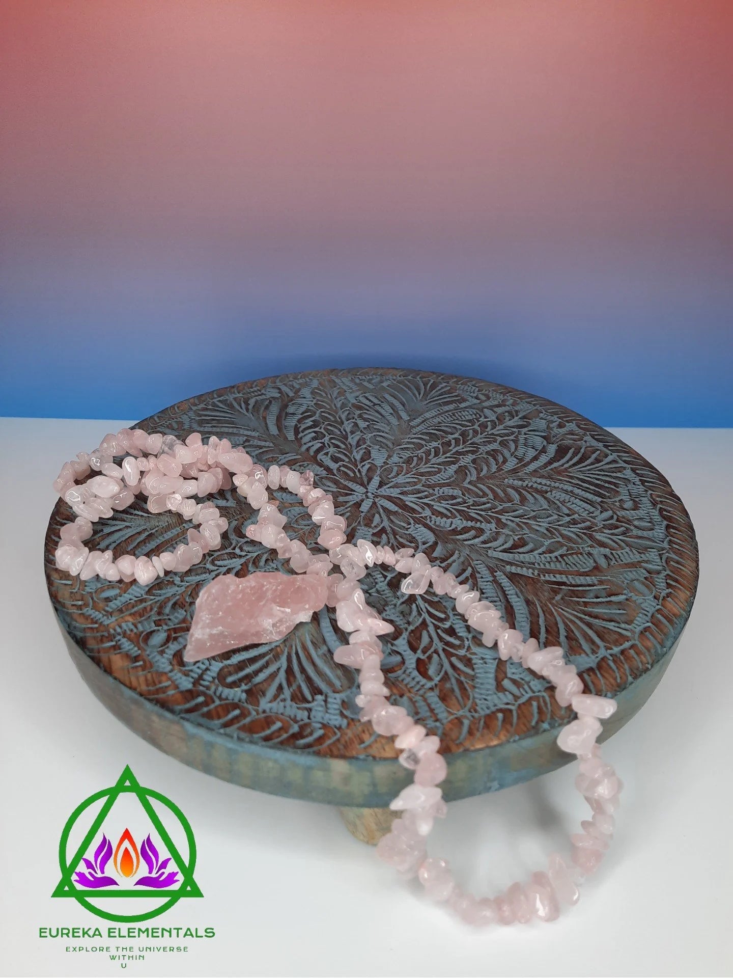 Rose Quartz Necklace and Crystal resting on a wooden stand with a red and blue ombre backdrop. 