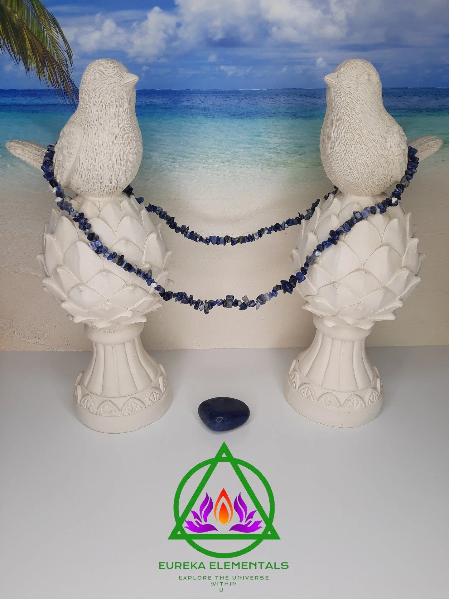 Sodalite Necklace and Crystal resting between two white dove statues with a beach backdrop.